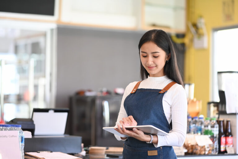 Young woman coffee shop owner wearing apron holding digital tablet ready to receive orders.
